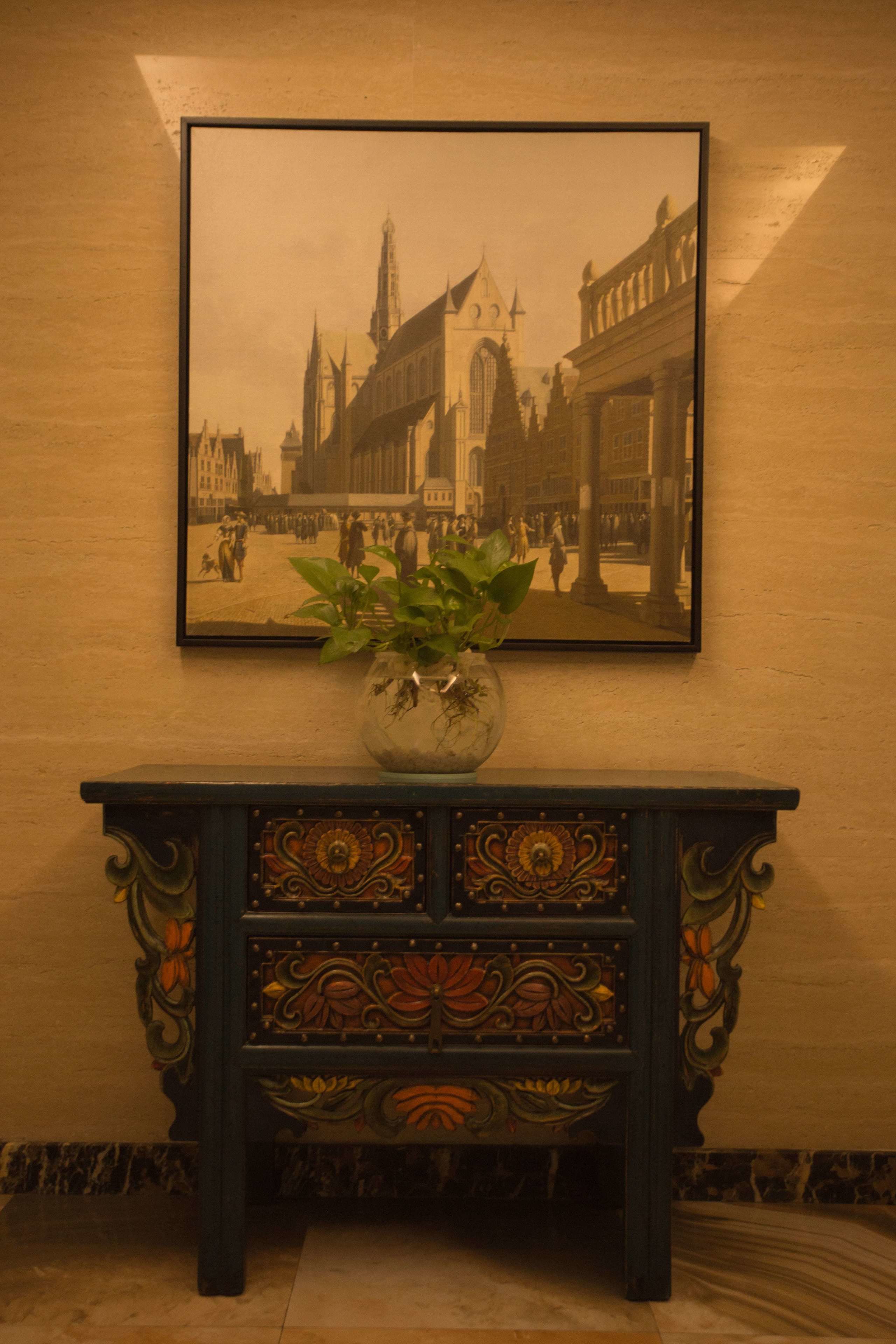 A framed historical photograph of a town square with a decorative console table below it, featuring intricate carvings and a potted plant.