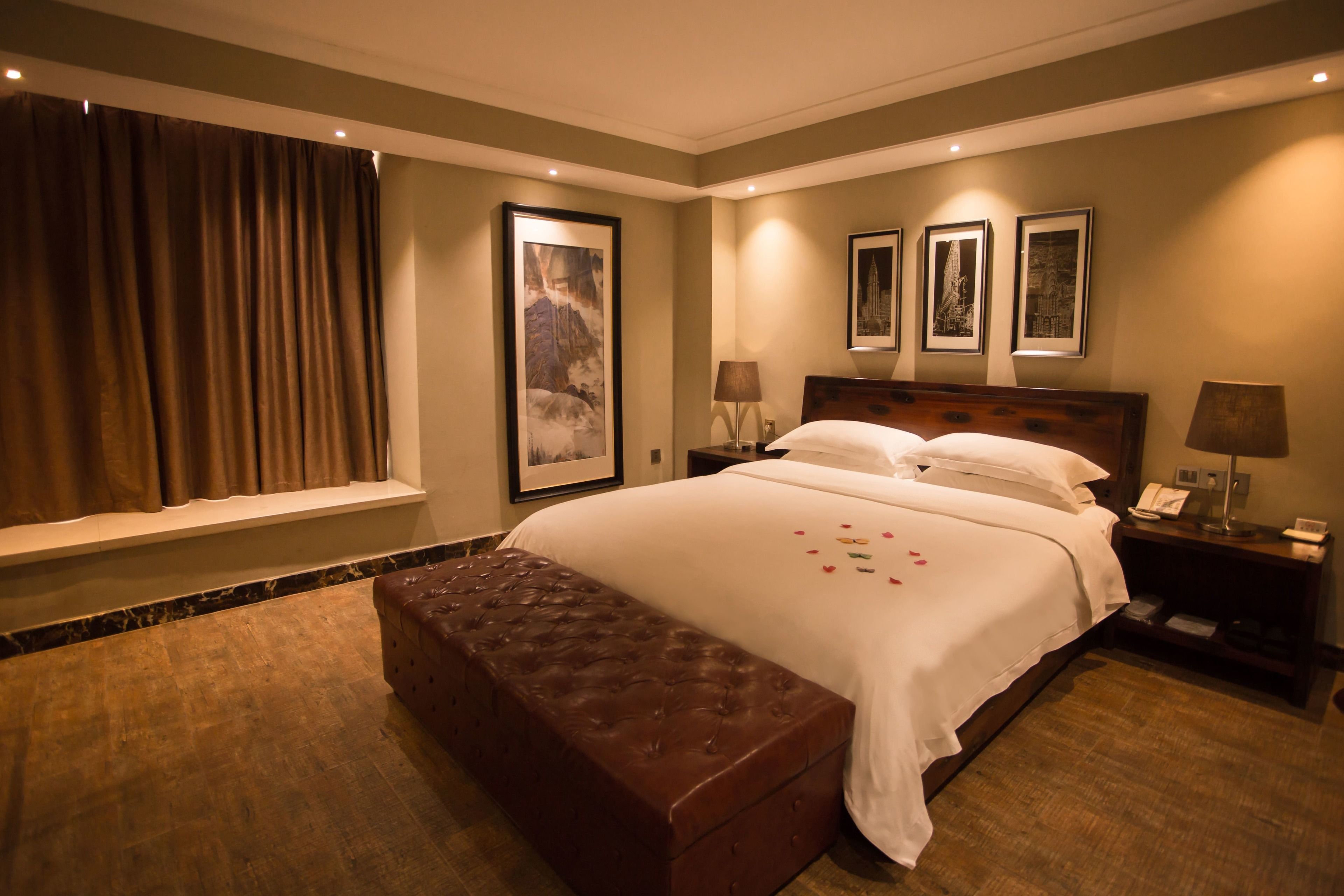 A hotel room with a large bed, a wooden bench, a window with curtains, and framed pictures on the wall.