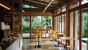 A cafe with wooden tables and chairs, a counter with food items, and large windows with a view of greenery. - SUNKISS pool.bed.cafe (Bangkok)