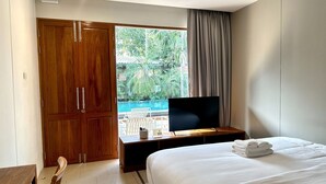 A hotel room with a bed, a TV on a wooden stand, a wooden wardrobe, a window with a view of a swimming pool and greenery, and a door. - SUNKISS pool.bed.cafe (Bangkok)