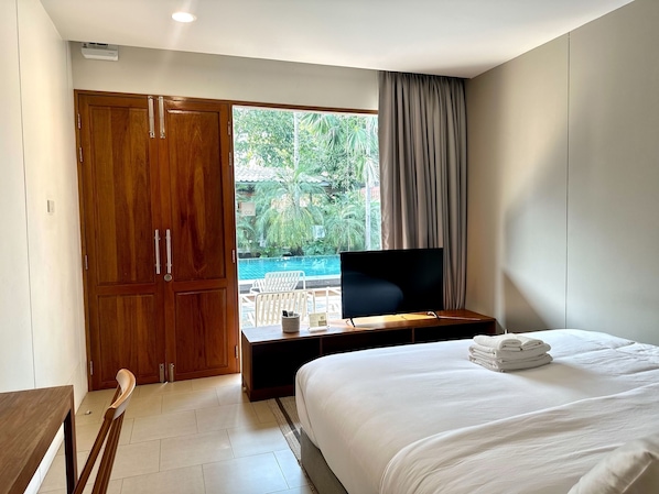 A hotel room with a bed, a TV on a wooden stand, a wooden wardrobe, a window with a view of a swimming pool and greenery, and a door. - SUNKISS pool.bed.cafe (Bangkok)