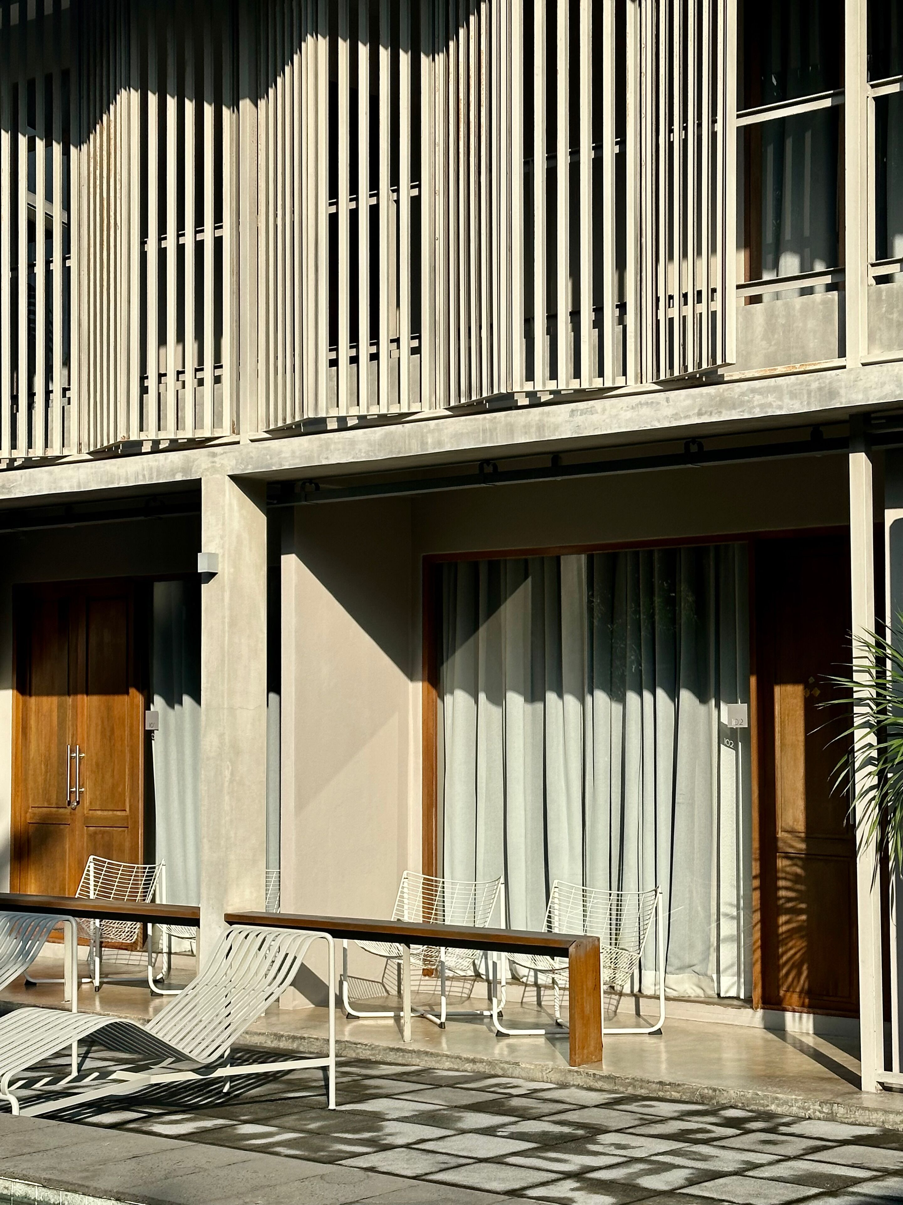 A modern building with a balcony, a wooden door, and a seating area with chairs and a bench.