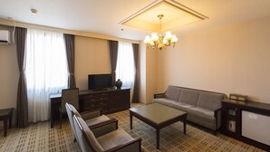 Habitación de hotel con sofá, sillones, una mesa de centro, televisor, un mueble bajo y una lámpara en el techo.