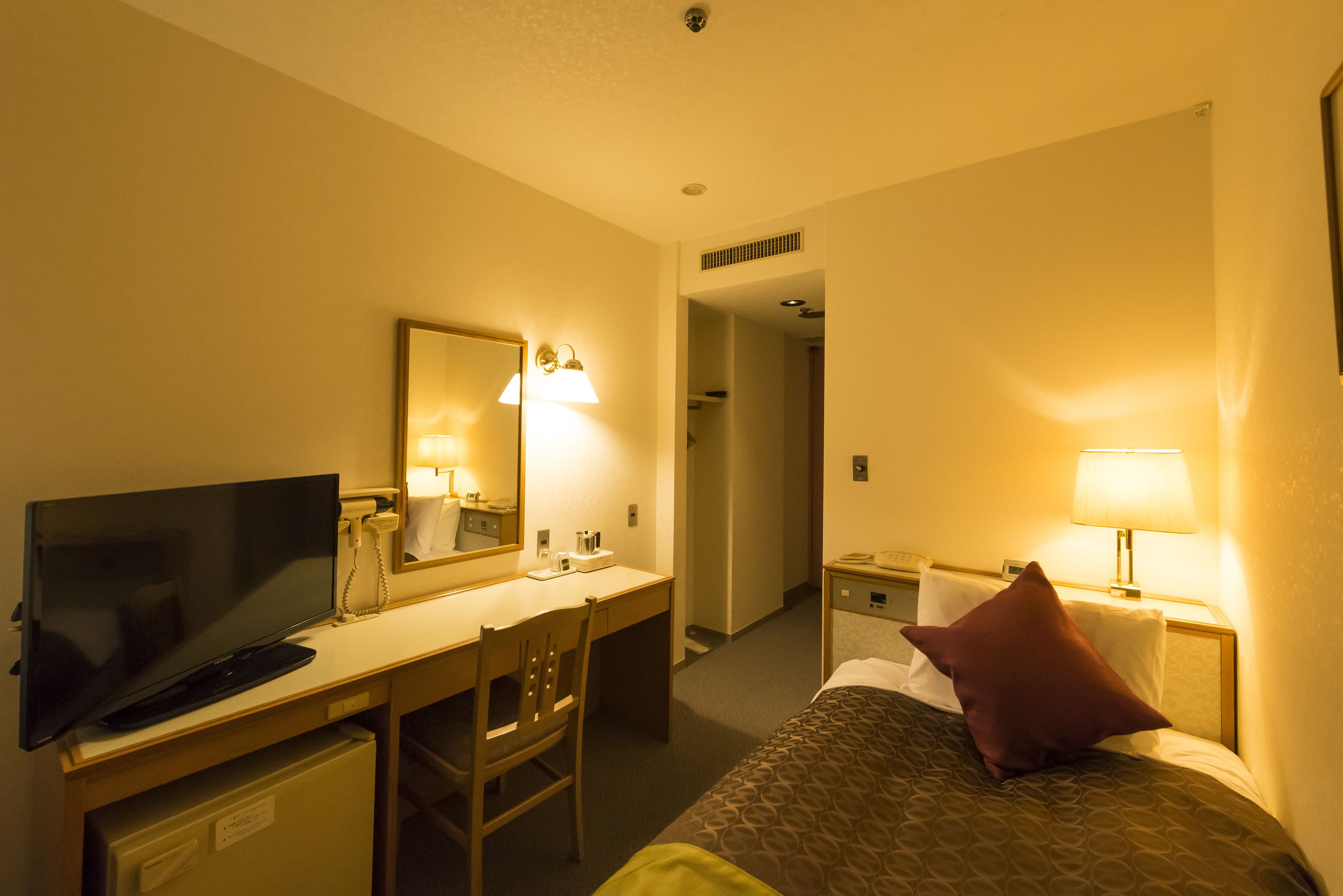 Una habitación de hotel con una cama, un escritorio con televisor, una silla, un espejo y una mesita de noche con lámpara.