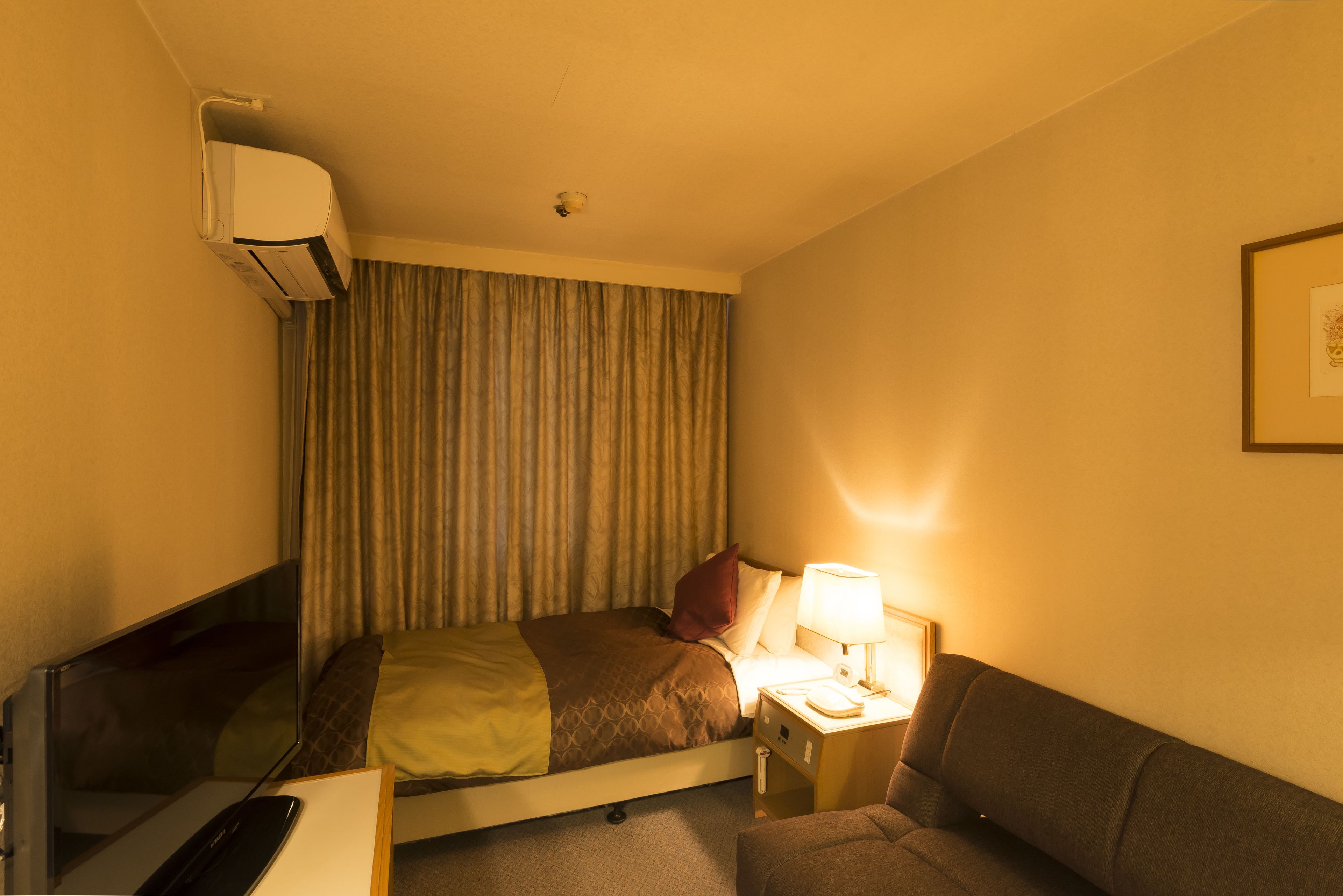 Ein Hotelzimmer mit einem Bett, einem Nachttisch mit Lampe, einem Fernseher, einem Sofa und einer Klimaanlage.