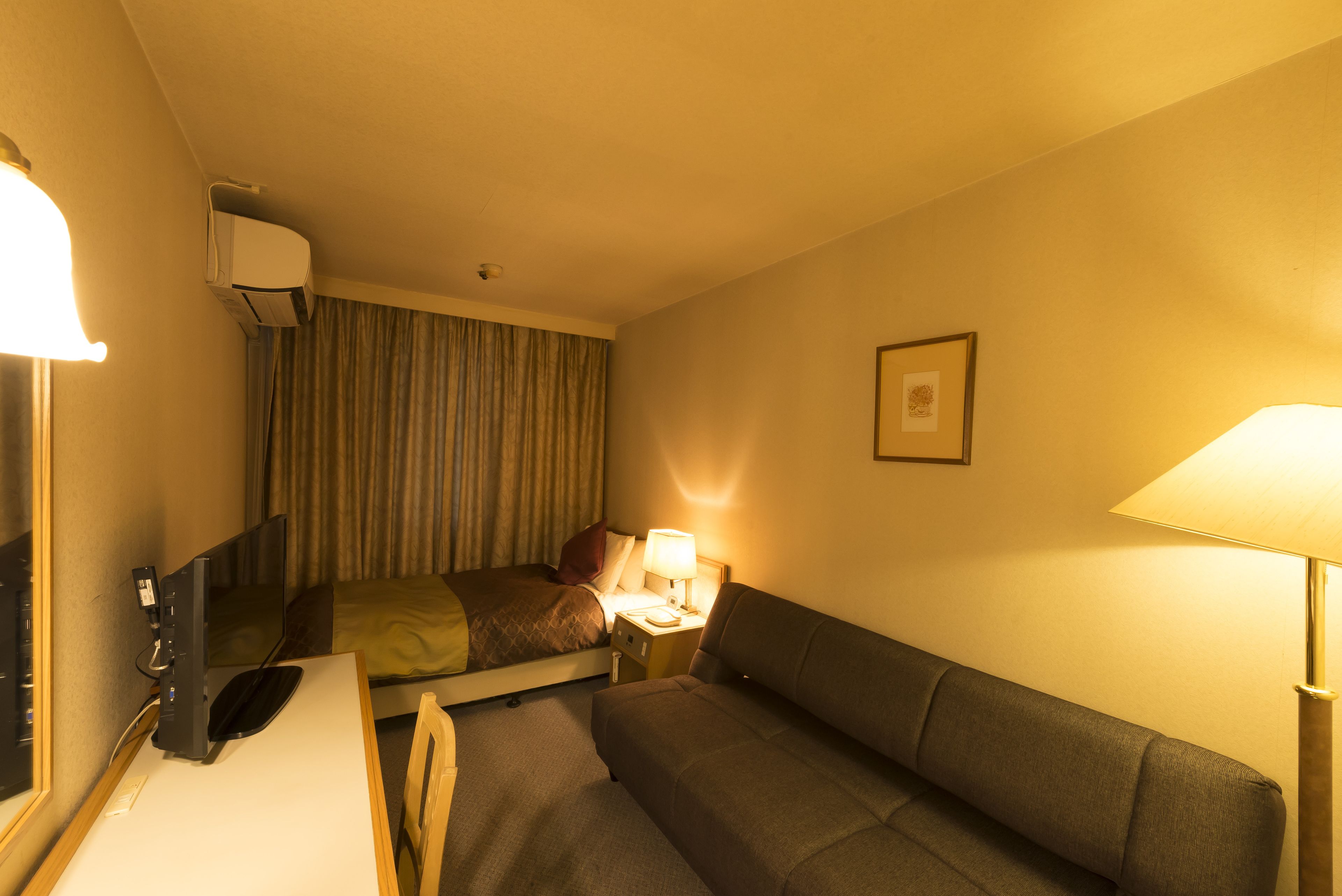 A hotel room with a bed, a desk with a computer, a couch, and a bedside table with a lamp.