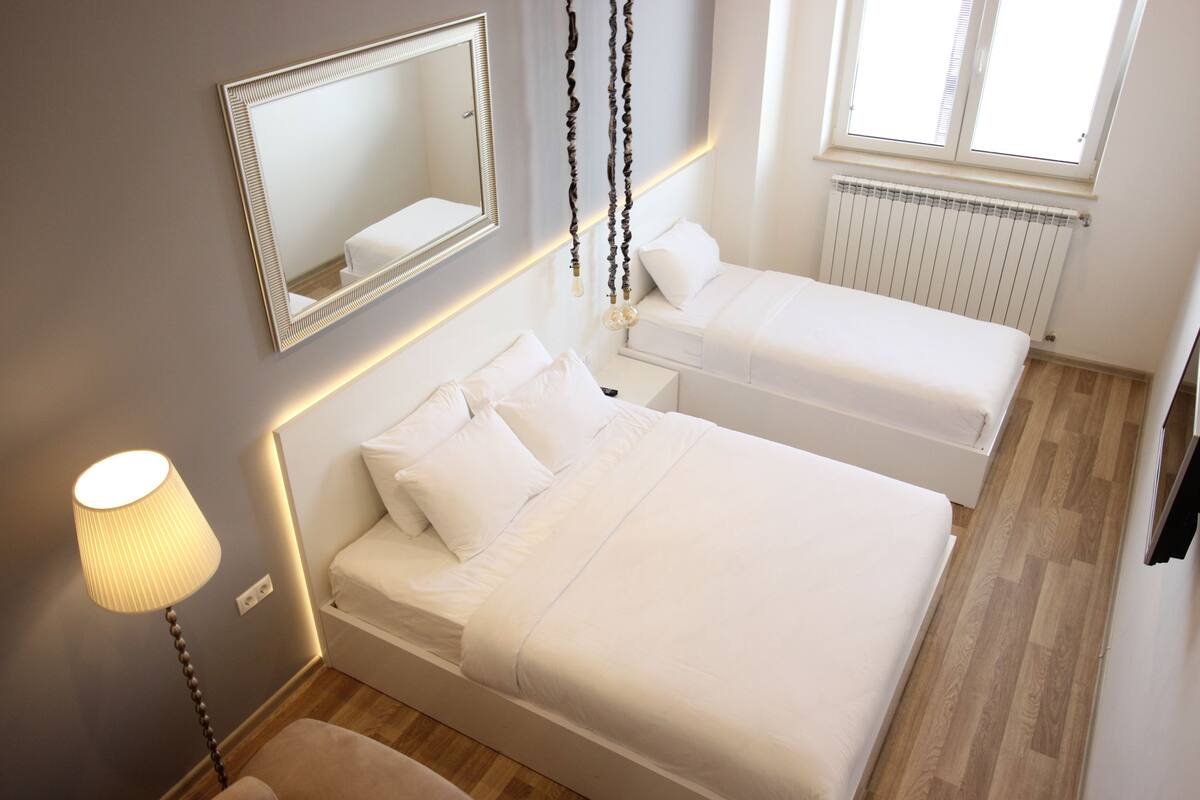 A bedroom with two beds, a bedside table, a lamp, a mirror, and a window with blinds.