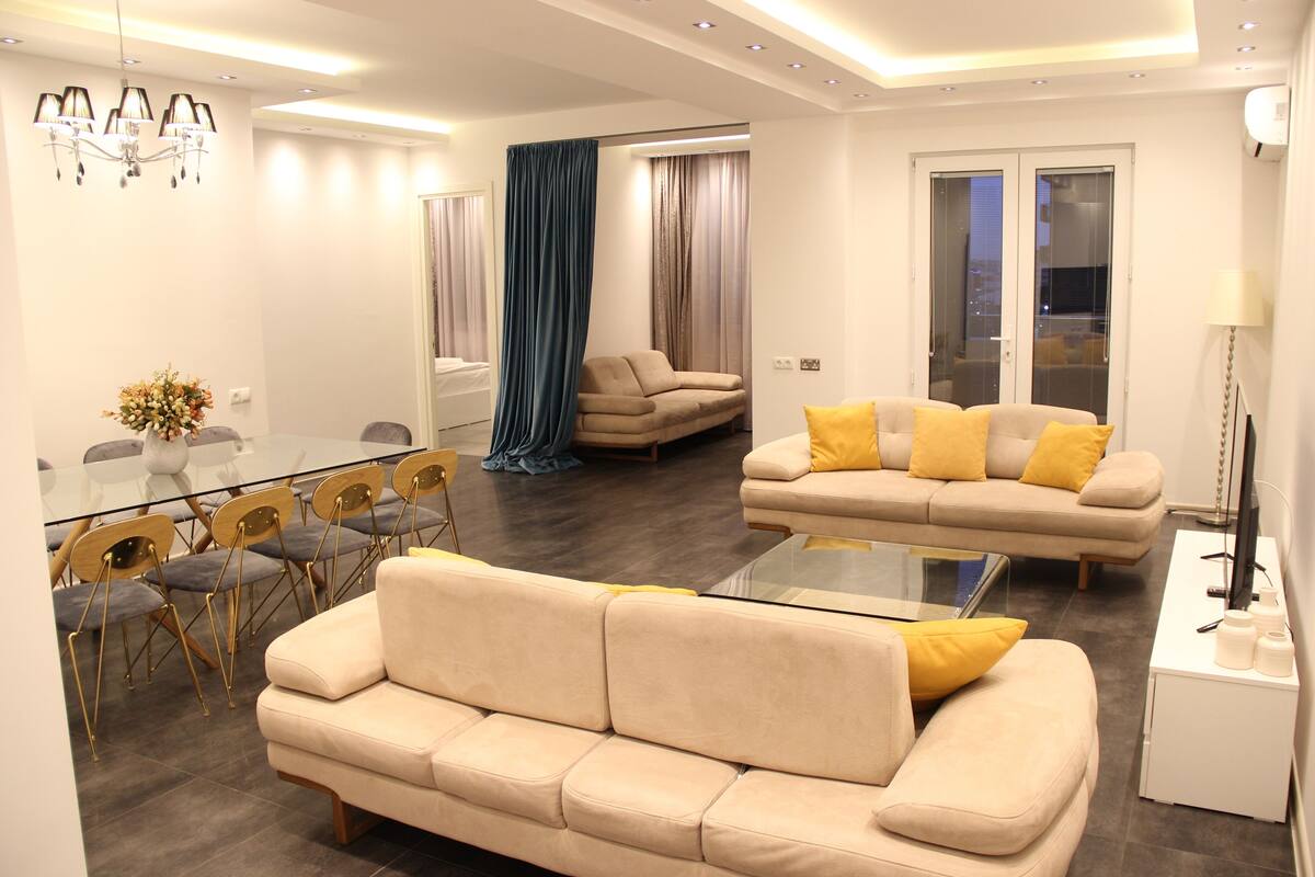 A modern living room with a beige sofa, yellow cushions, a glass coffee table, and a dining area with a glass table and chairs.