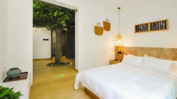 Une chambre à coucher avec un lit, une table de chevet, une lampe fixée au mur, une tête de lit en bois et un arbre en arrière-plan.