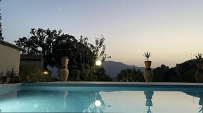 A swimming pool with clear water, surrounded by potted plants and trees, with a mountain in the background. - Le due querce (Pettineo)