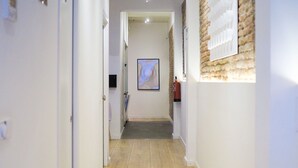 A hallway with a wooden floor, white walls, and a brick accent wall. There is a door on the left and a framed picture on the right. - Arte Suites by Olala Homes (L'Hospitalet de Llobregat)