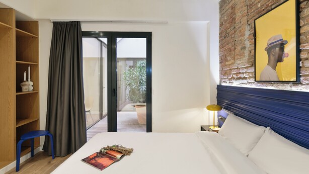 A hotel room with a bed, a wooden shelf, a blue stool, a curtain, a door leading to a balcony, and a framed picture on the wall. - Arte Suites by Olala Homes (L'Hospitalet de Llobregat)