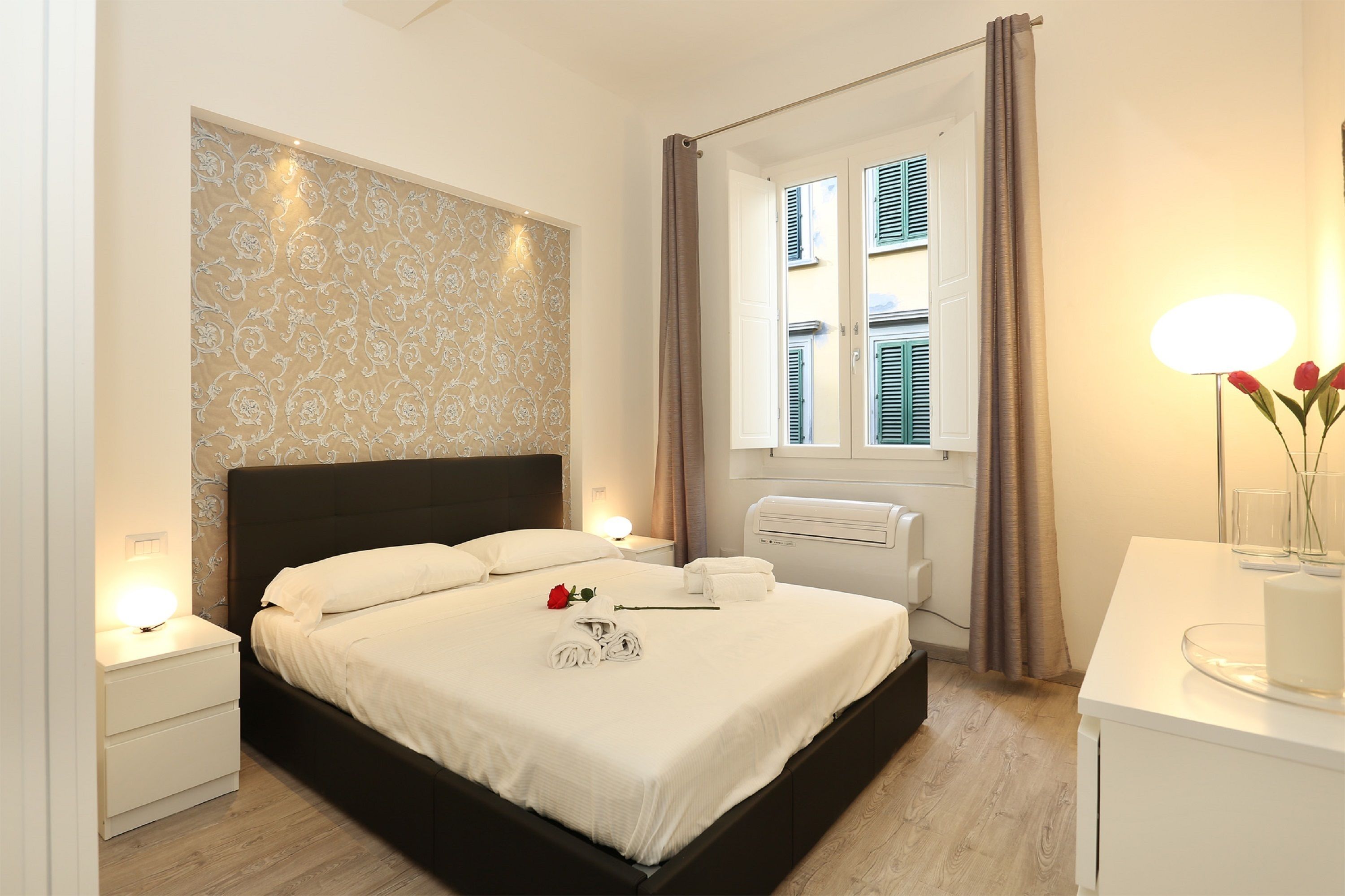 A neatly made bed with white linens, a bedside lamp, and a vase with red flowers on a white nightstand.