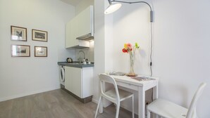 Une petite cuisine équipée d’un évier, d’une bouilloire et d’une machine à café. Trois tableaux encadrés ornent le mur. Une table avec un vase de fleurs et un magazine se trouve au premier plan.