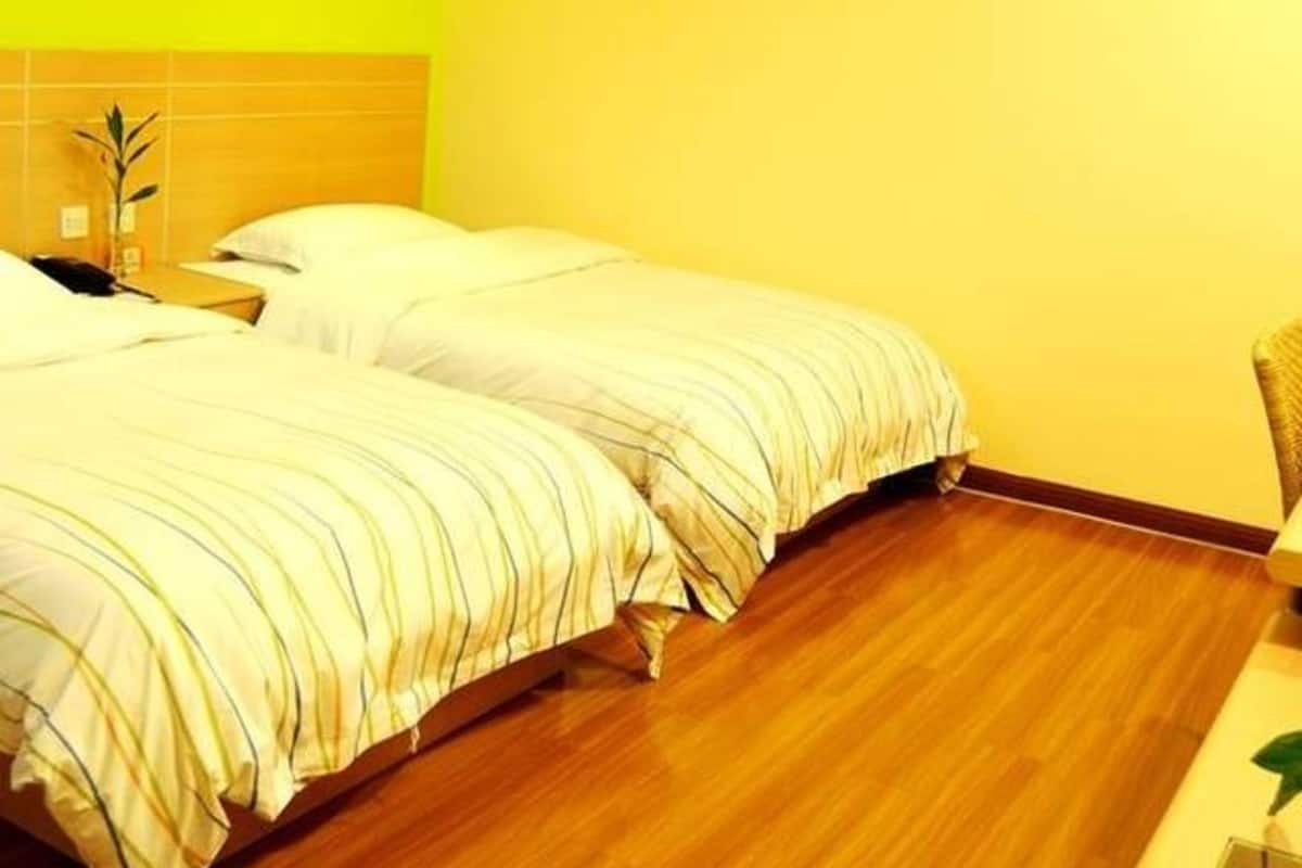 Ein Hotelzimmer mit zwei Betten, Holzboden, einer leuchtend gelben Wand und einer kleinen Pflanze auf dem Nachttisch.