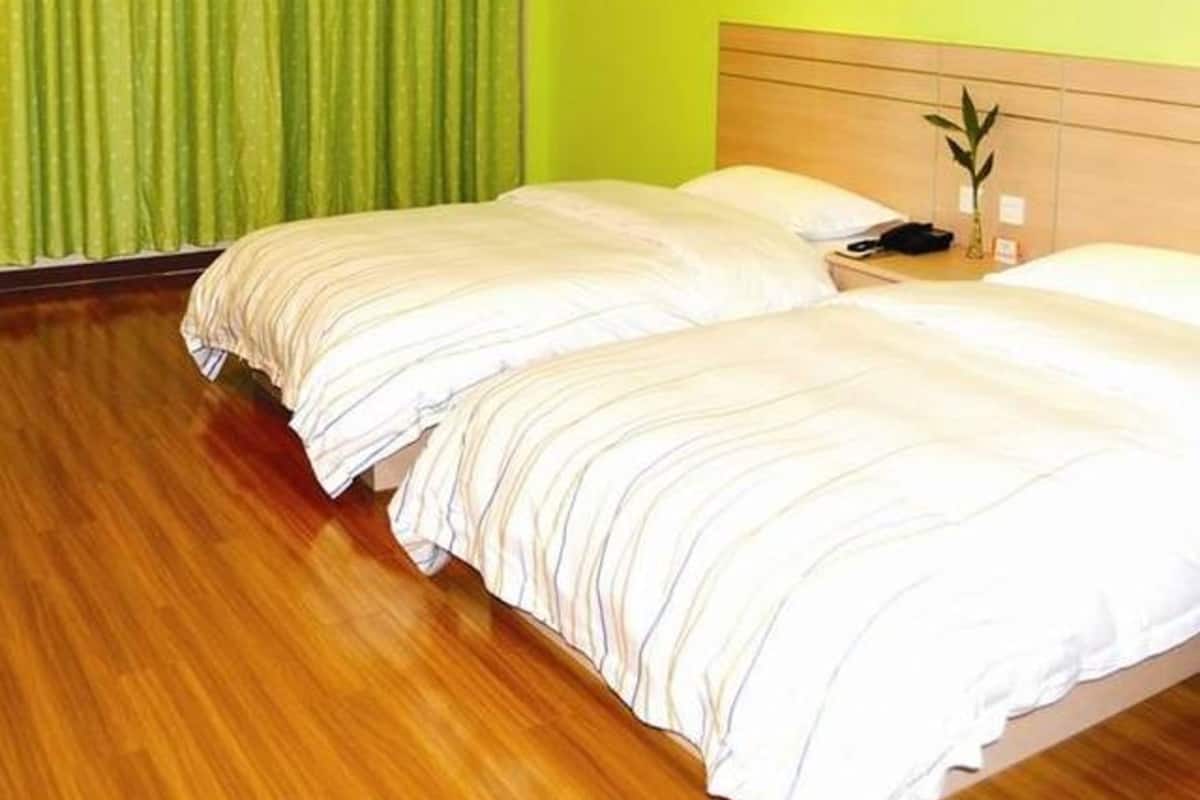 Ein Hotelzimmer mit zwei Betten, grünen Vorhängen, Holzboden und einer kleinen Pflanze auf dem Nachttisch.