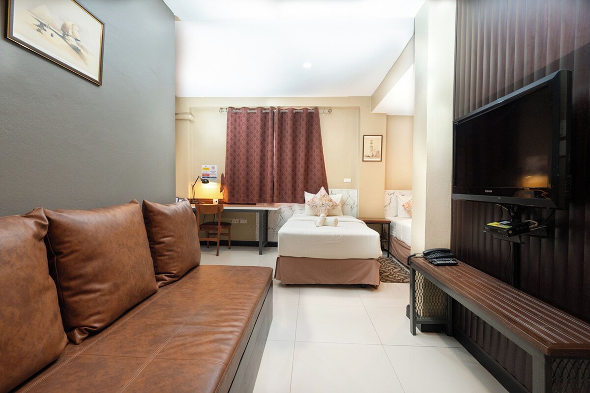 A hotel room with a brown leather sofa, a small bed with a white mattress, a desk with a chair, a television, and a shower curtain.