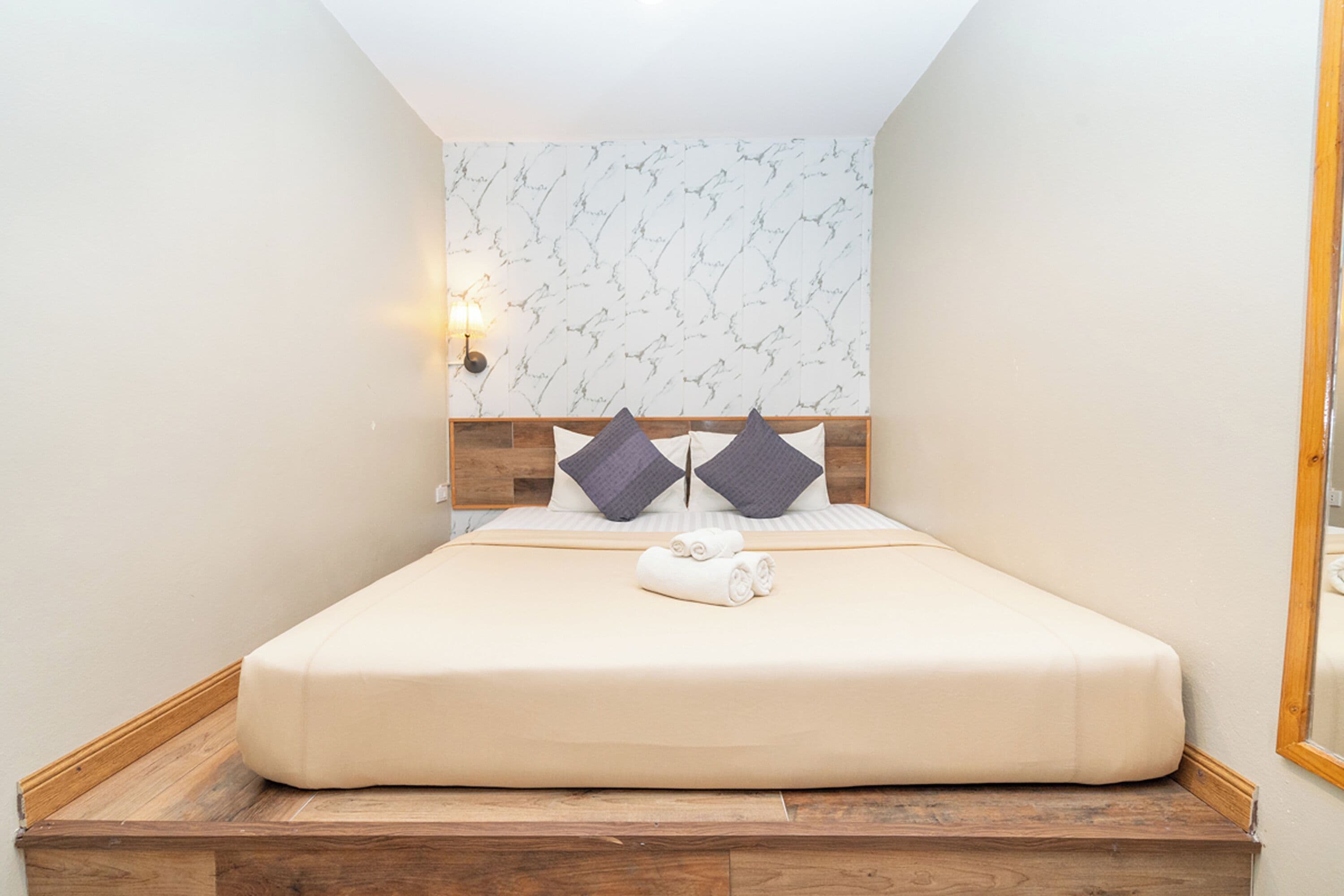 A neatly made bed with a wooden headboard, a wall-mounted light, and a marble-patterned wall.