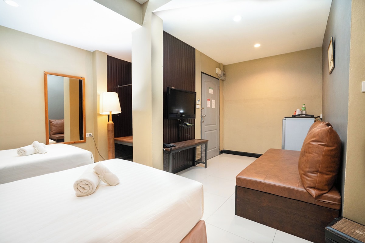 A hotel room with two beds, a TV, a bench, and a small kitchenette.