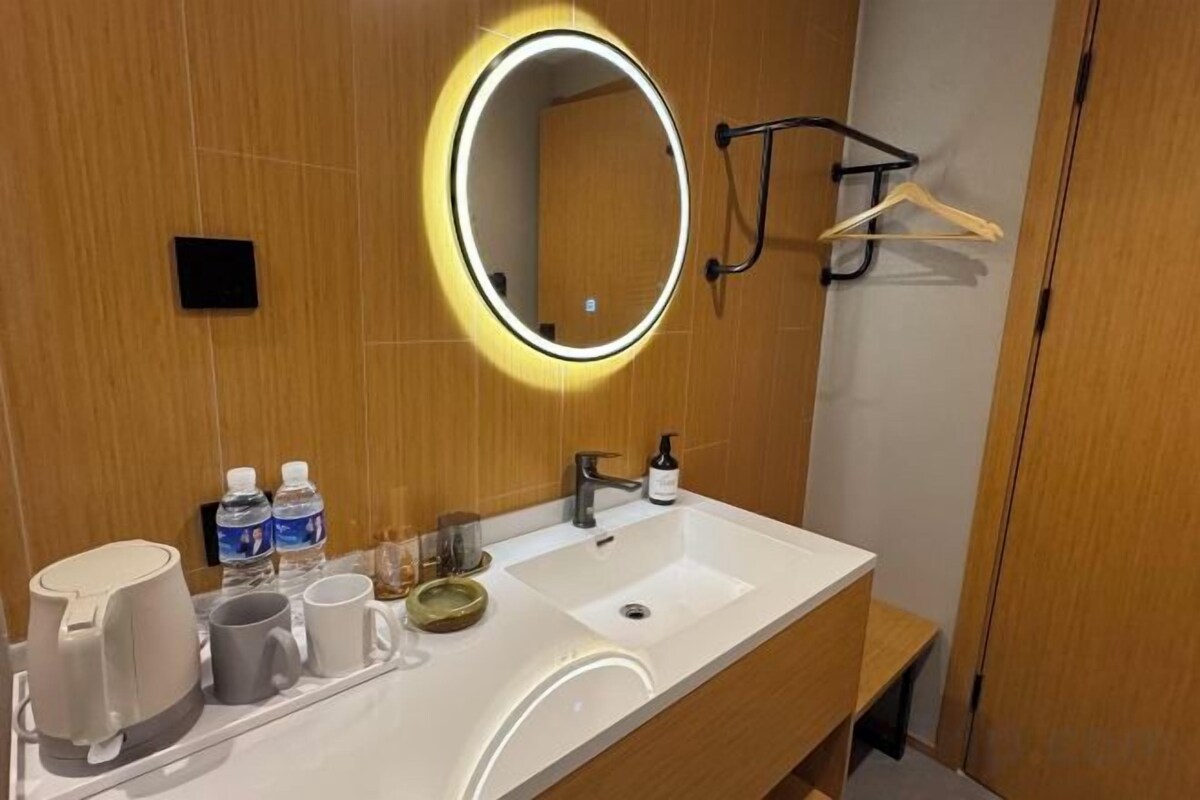 Une salle de bain d’hôtel avec un lavabo blanc, un miroir rond avec un éclairage intégré et un porte-serviettes.