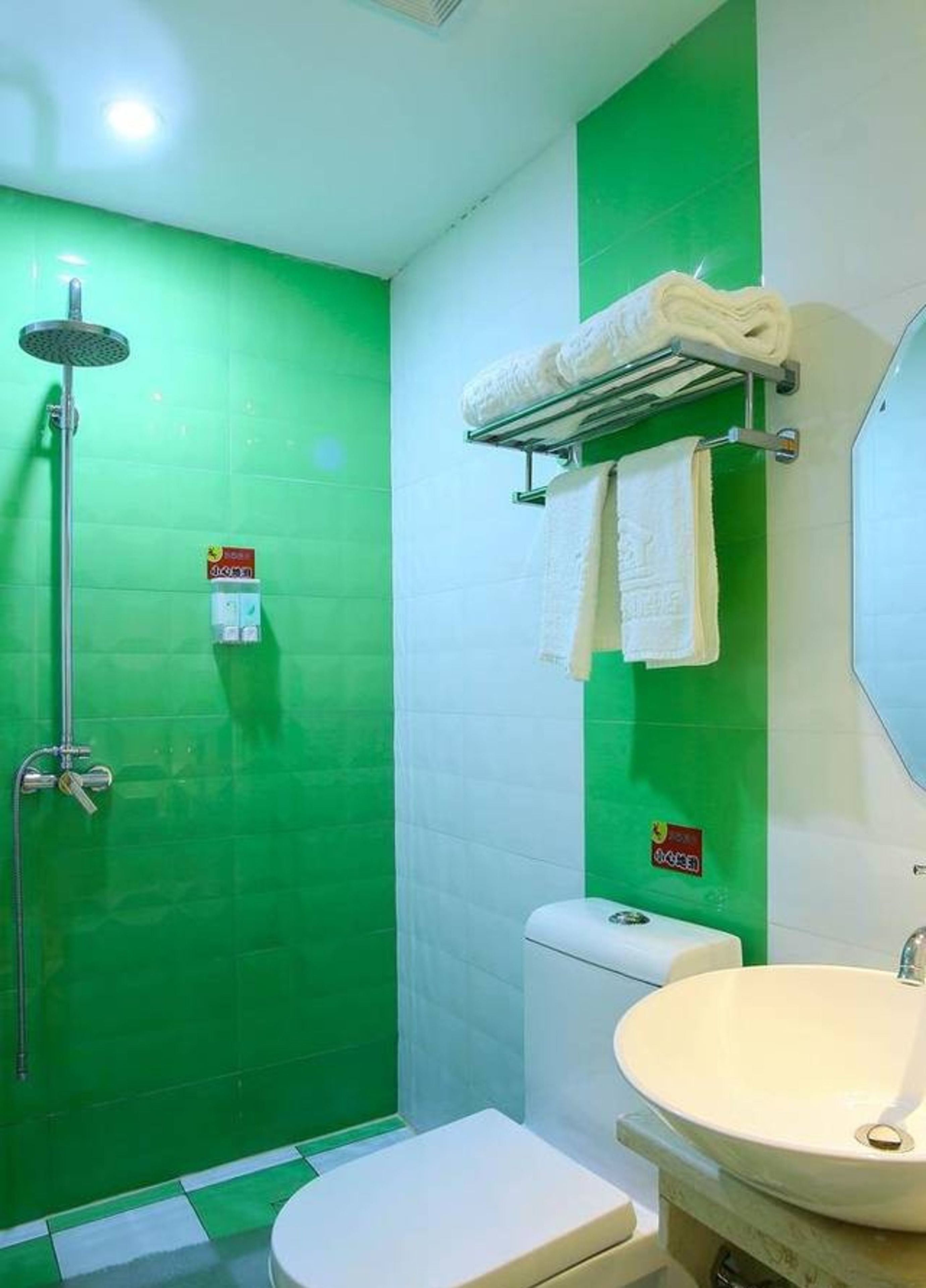 A bathroom with a green wall, a white toilet, a shower with a handheld showerhead, a towel rack with a towel, and a sink with a mirror above it.