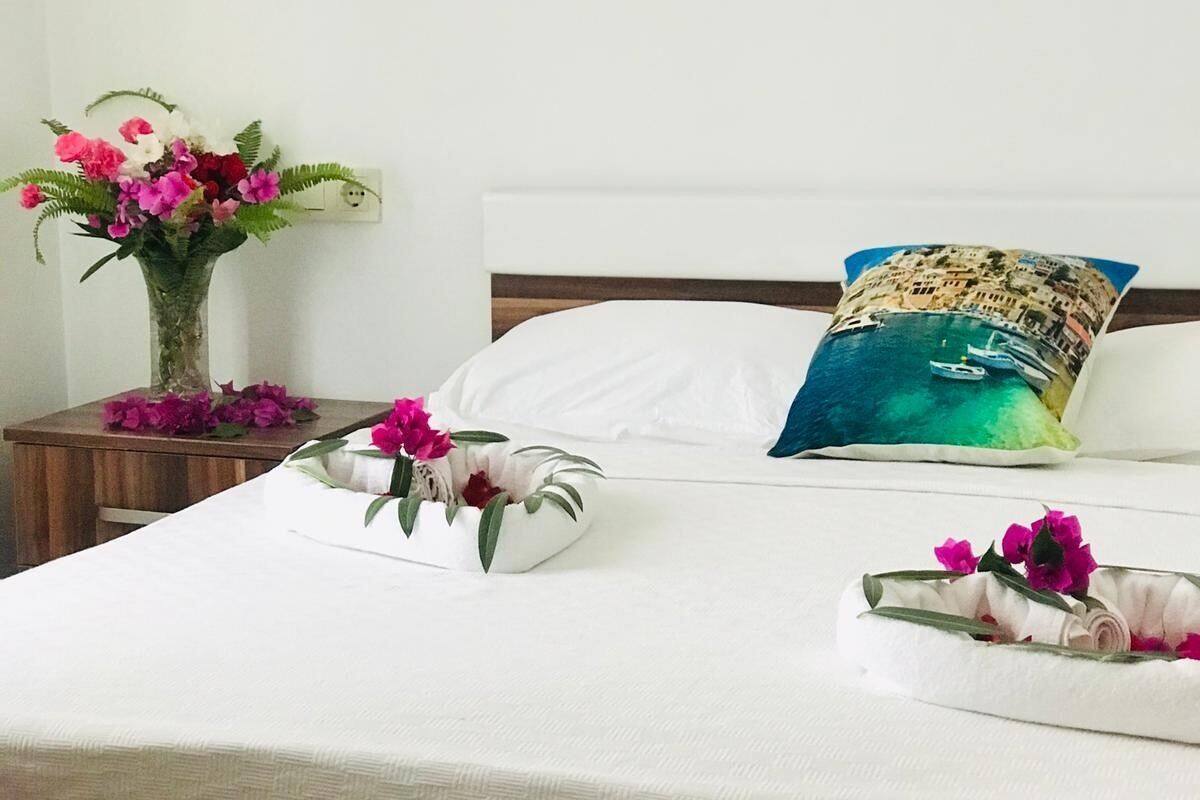 A neatly made bed with a white bedspread, a pillow with a coastal scene, and a vase of flowers on a bedside table.
