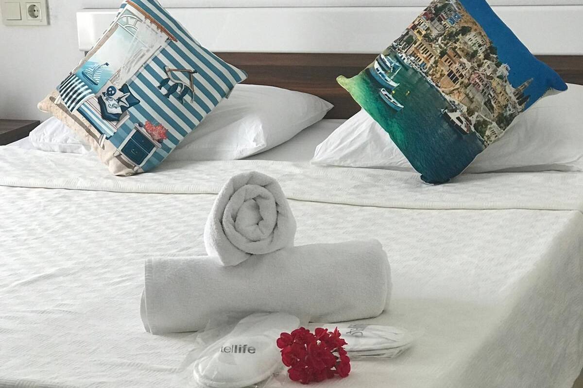 A neatly made bed with white bedding, a rolled white towel, and a small red flower.