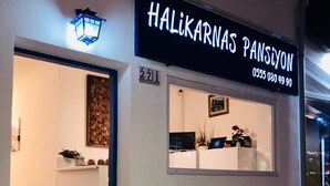 A hotel entrance with a reception desk, a signboard with the name 'HALIKARNAS PANSION' and a phone number, and a patterned tile floor. - Halikarnas Pansiyon (Bodrum)