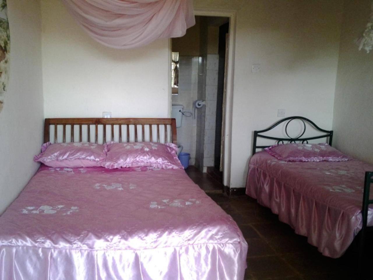 Un dormitorio con dos camas, cada una con una colcha rosa y almohadas a juego. Una cama tiene un cabecero de madera y la otra, un cabecero de metal.  Una cortina rosa cuelga del techo, y se ve un cubo azul en el suelo.
