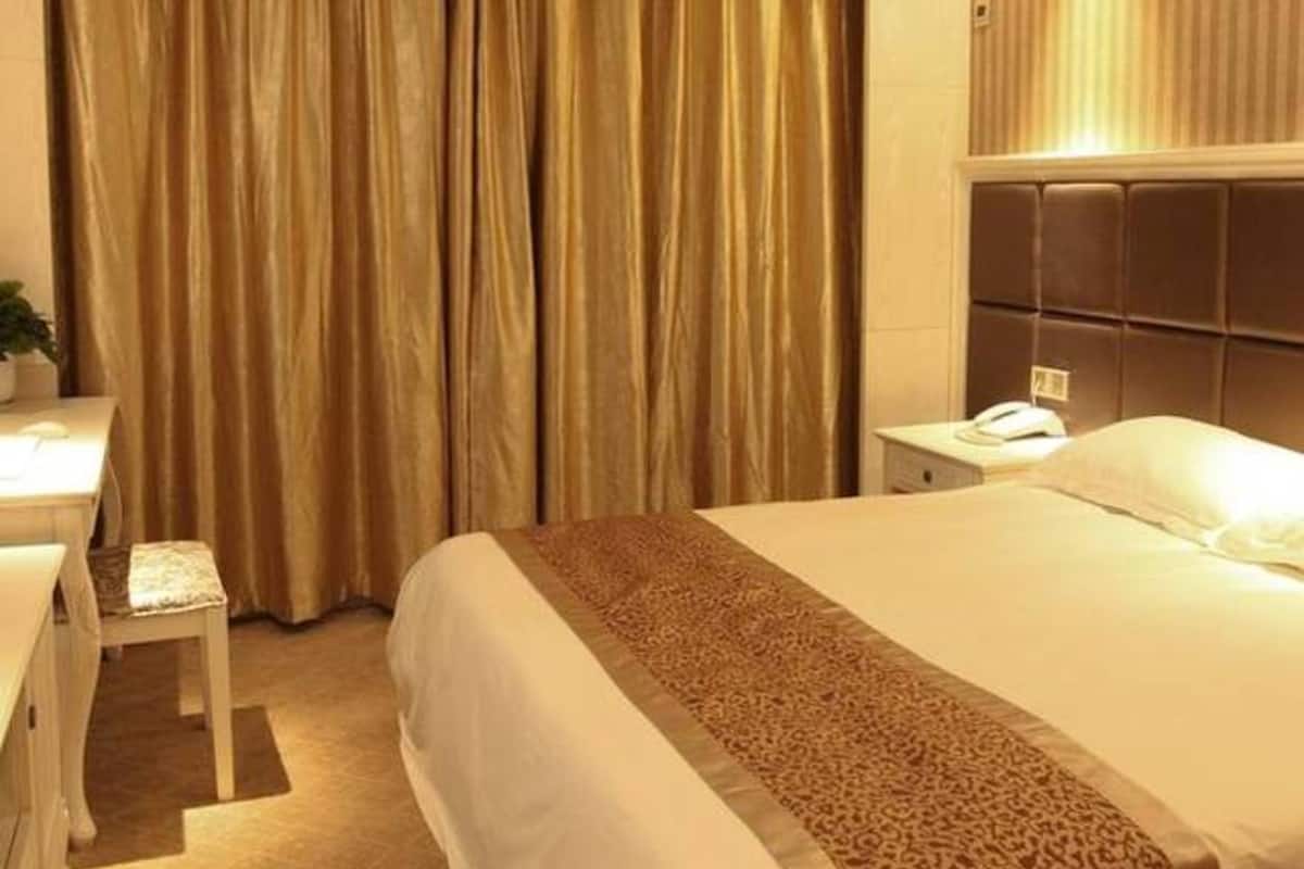 A hotel room with a large bed, a desk with a chair, a bedside table, and a large window with curtains.