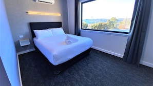 Habitación de hotel con una cama bien hecha, un ventanal con vistas, un equipo de aire acondicionado y un estante en la pared.