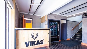 Un área de recepción con un mostrador de madera que muestra el logotipo de 'VIKAS', una escalera y un letrero que indica un bar restaurante en el piso superior.