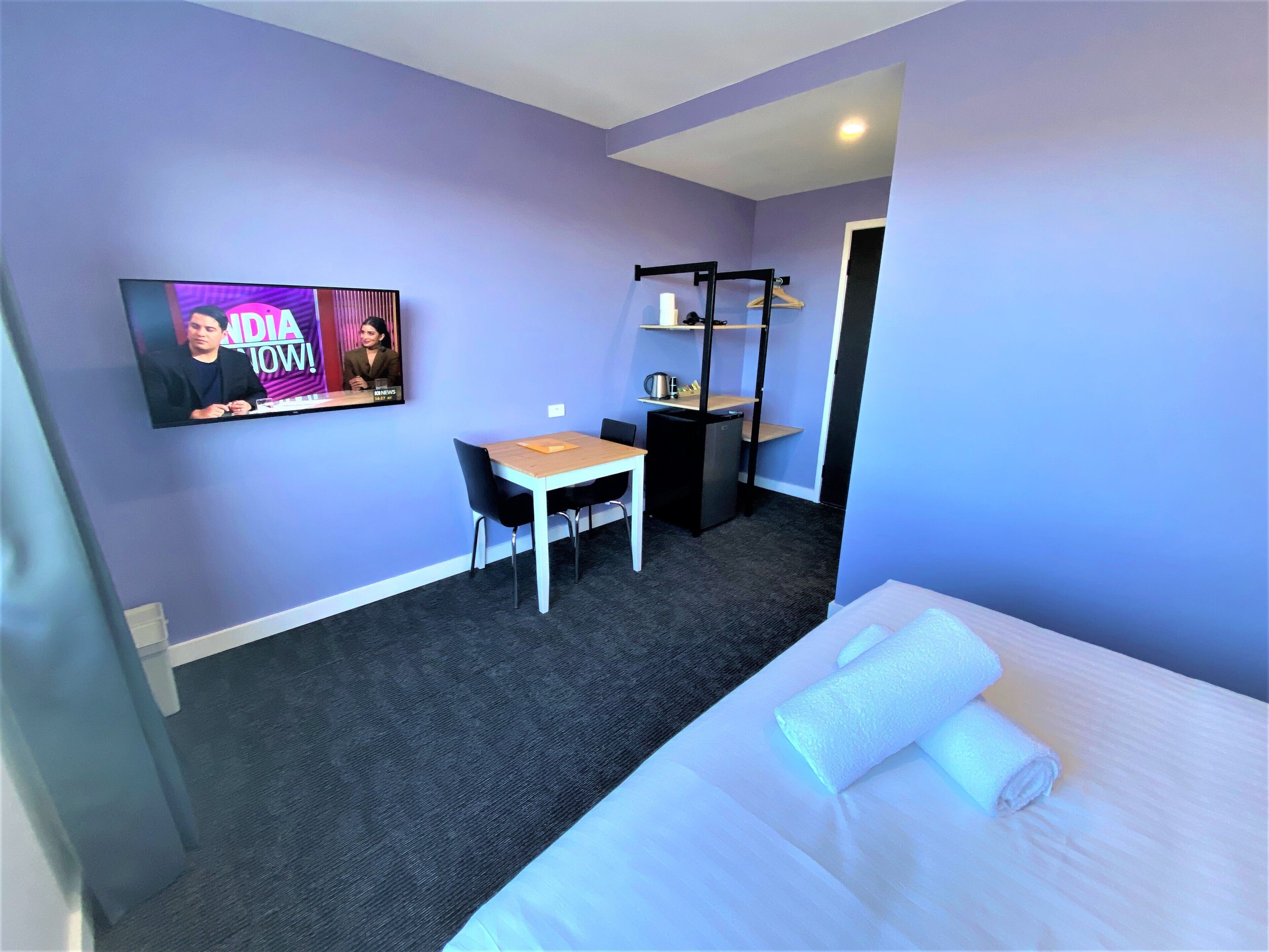 Een hotelkamer met een bed, een bureau, een stoel, een aan de muur bevestigde televisie en een klein kitchenette.