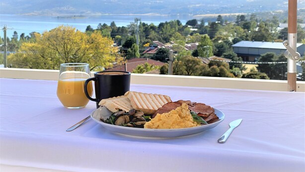 Un desayuno con un plato de comida, un vaso de jugo de naranja y una taza negra sobre una mesa con vistas a un lago y montañas.