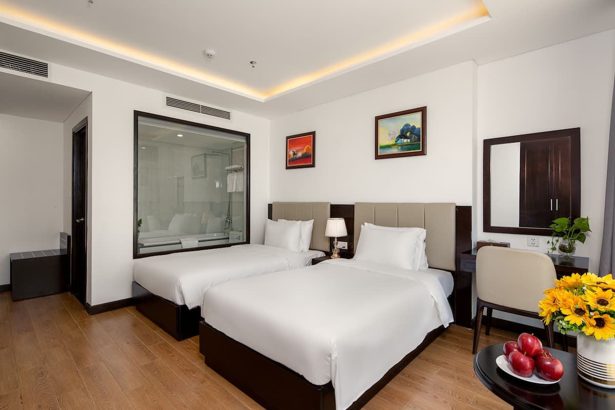 Habitación de hotel con dos camas, un ventanal grande, un cuadro, y una mesa con fruta y un jarrón de flores.