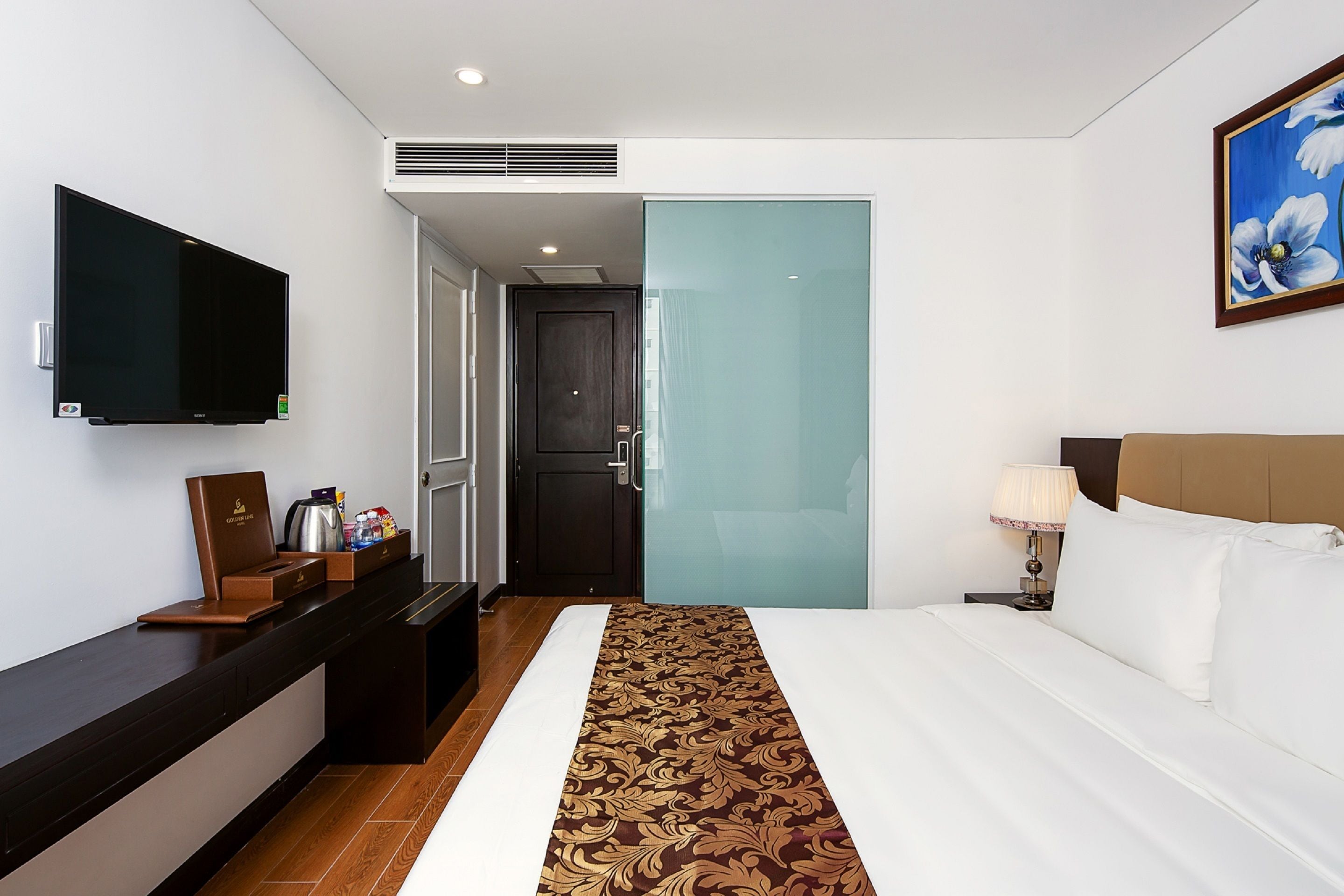 A hotel room with a bed, a TV, a desk with a coffee maker, and a painting on the wall.
