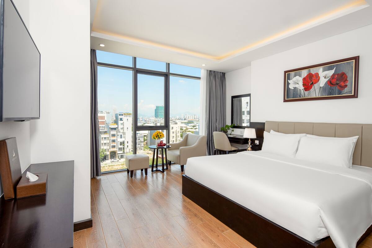 Habitación de hotel con una cama grande, un escritorio, una silla, un televisor y un amplio ventanal con vistas a la ciudad.