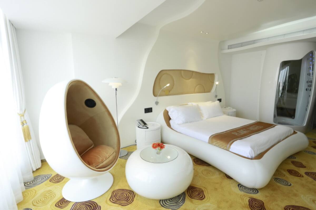 Una habitación de hotel moderna con techo curvo, una silla de diseño con forma de huevo, una cama con cabecera, una mesita de noche y un espejo grande.