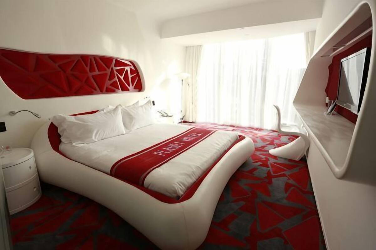 Una habitación de hotel moderna con una cama curvada, decoración en colores rojo y blanco, un ventanal con cortinas y un televisor de pantalla plana.