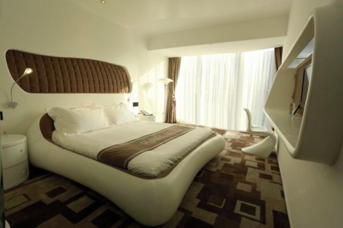 A modern hotel room with a curved bed, a large window with curtains, a flat-screen TV, and a bedside table with a lamp.