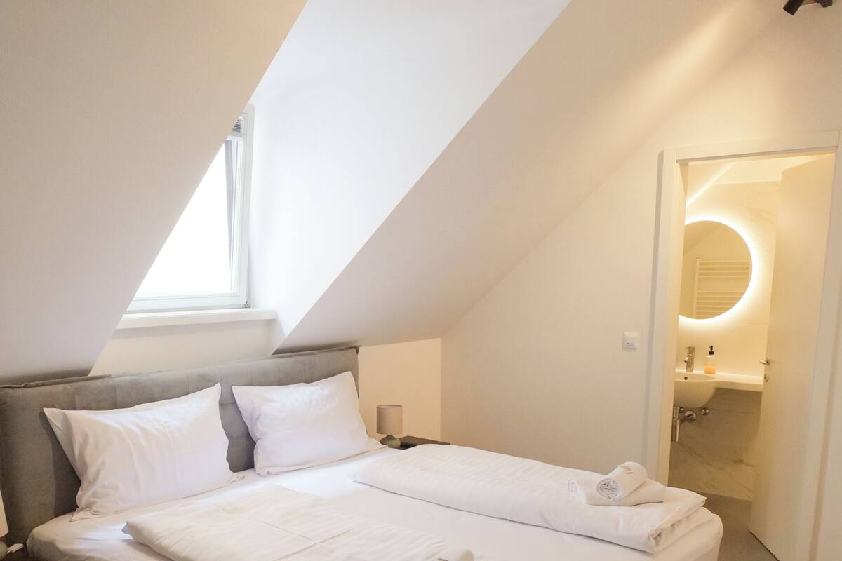 Un dormitorio con techo inclinado, una cama con ropa de cama blanca, una mesita de noche y un buró.