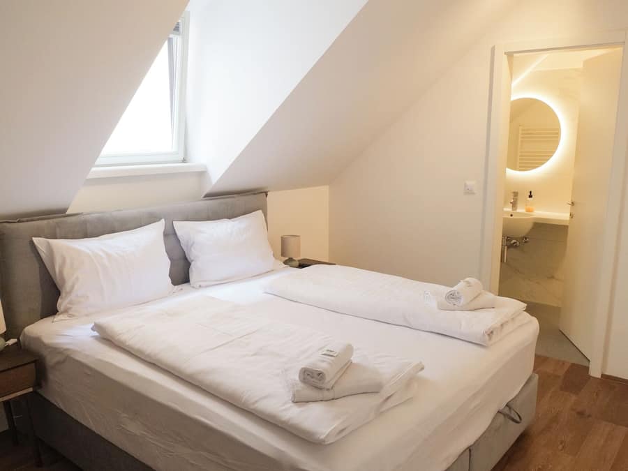 Une chambre avec un plafond en pente, un lit recouvert de draps blancs, une table de chevet et une commode.