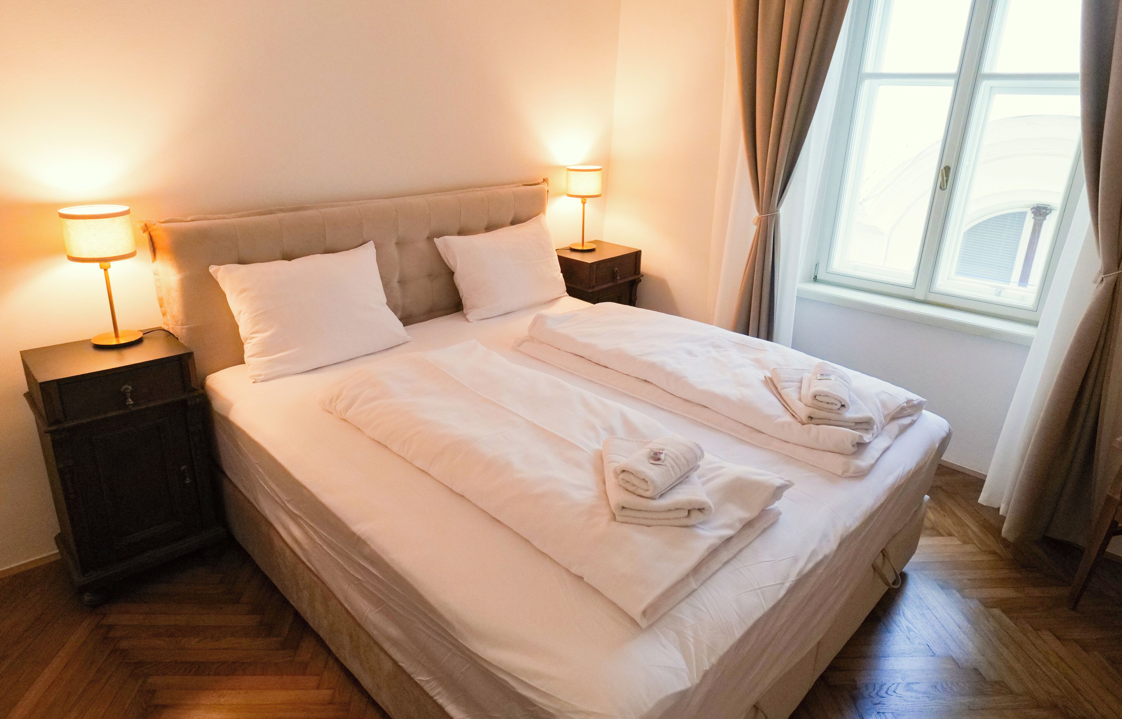 Uma cama de casal com lençóis brancos, duas luminárias de cabeceira e uma janela com cortinas.
