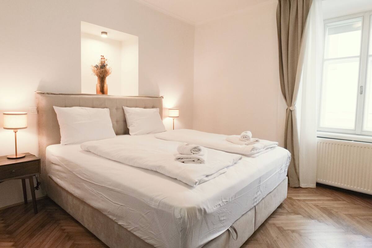 Una cama bien hecha con sábanas y toallas blancas, una mesita de noche con una lámpara y una ventana con cortinas.