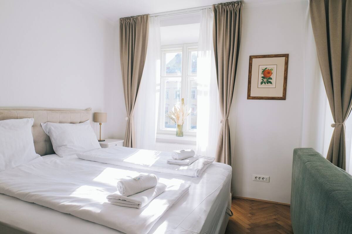 Habitación de hotel con una cama grande, un sillón verde, una ventana con cortinas y un cuadro enmarcado en la pared.