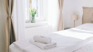 A neatly made bed with white linens and a rolled towel, a bedside lamp, and a window with curtains.