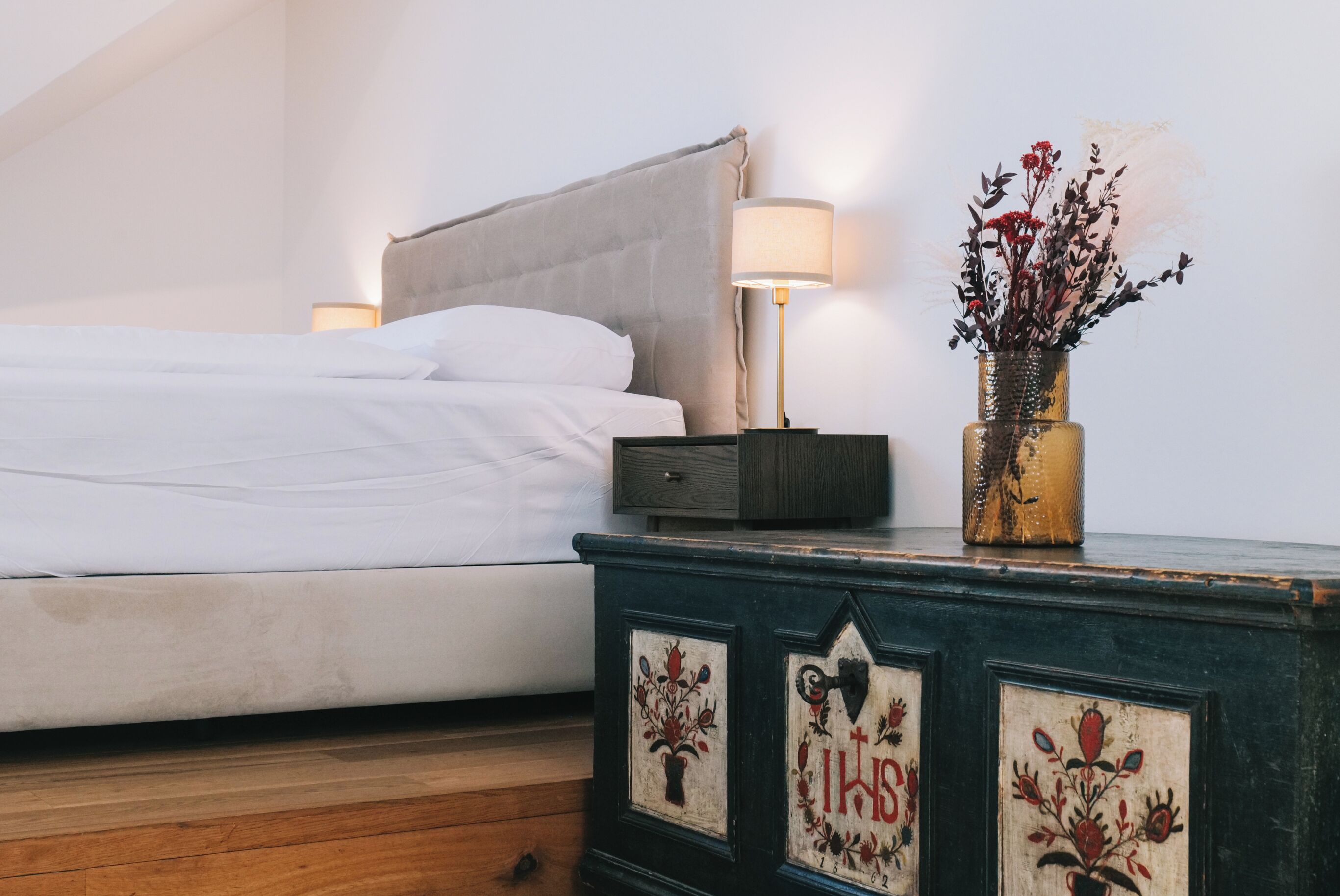 A neatly made bed with white linens, a bedside table with a lamp and a vase of flowers, and a dresser with floral designs.