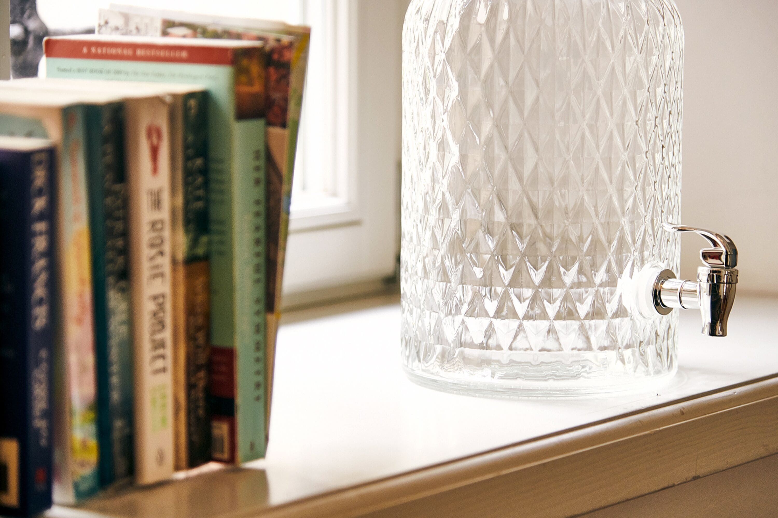 A clear glass pitcher on a shelf with books behind it.
