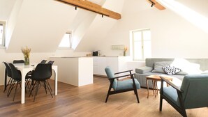 Un espacioso apartamento tipo loft con techo alto, vigas de madera a la vista, una zona de comedor con mesa blanca y sillas negras, un sofá gris y una pequeña cocina.