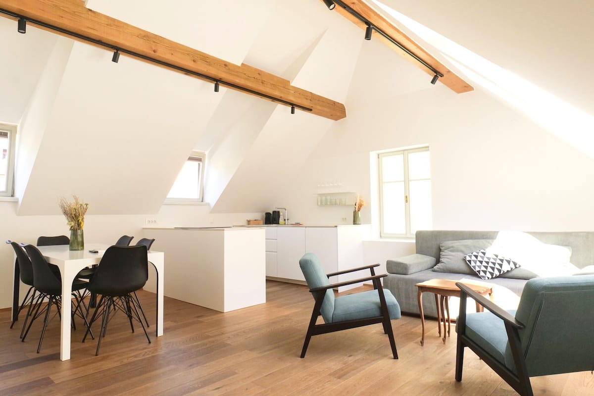 Un espacioso apartamento tipo loft con techo alto, vigas de madera a la vista, una zona de comedor con mesa blanca y sillas negras, un sofá gris y una pequeña cocina.